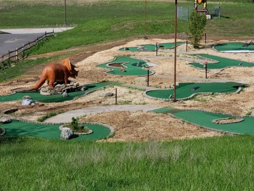 A mini-golf course featuring a dinosaur statue, surrounded by green turf and gravel paths.