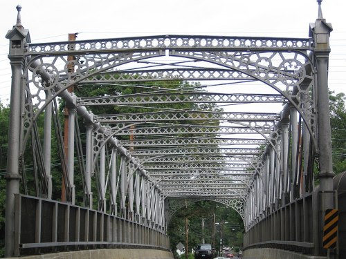 A decorative metal bridge with intricate latticework, spanning a road surrounded by trees and power lines.