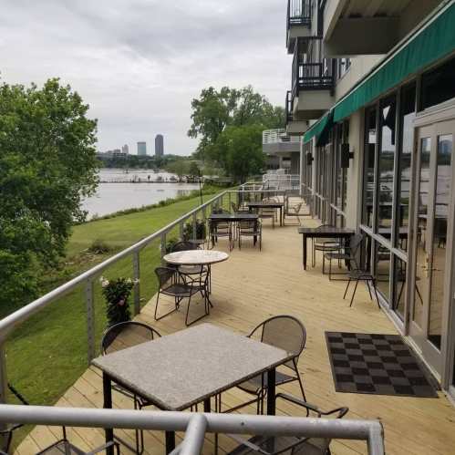 A riverside patio with empty tables and chairs, overlooking a green lawn and distant city skyline under a cloudy sky.