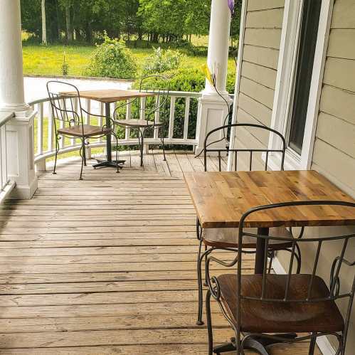 Two wooden tables with metal chairs on a porch, surrounded by greenery and a peaceful outdoor setting.
