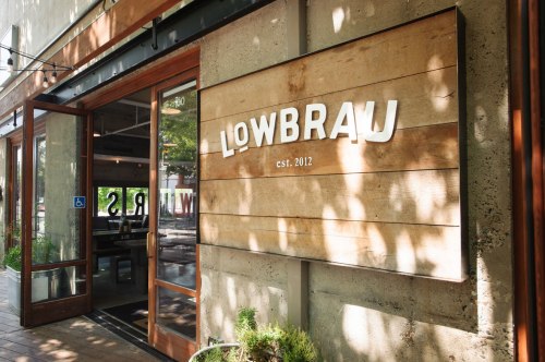 Exterior of Lowbrau restaurant with a wooden sign and large glass doors, surrounded by greenery. Established in 2012.