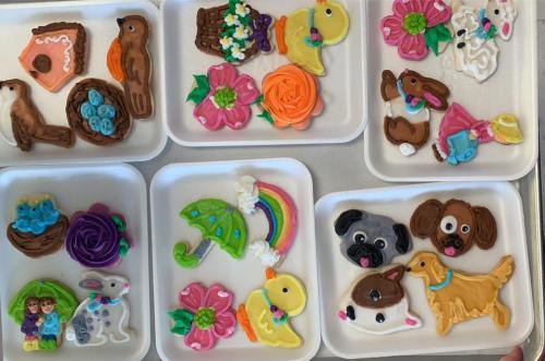 Colorful decorated cookies in various shapes, including animals, flowers, and a rainbow, arranged on white trays.