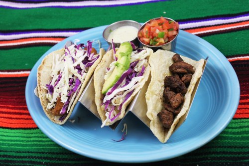 Three tacos on a blue plate, filled with cabbage, avocado, and seasoned meat, served with salsa and sauce on the side.