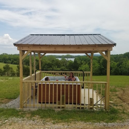 A wooden gazebo with a hot tub, surrounded by green fields and trees, under a partly cloudy sky. Two people relax inside.