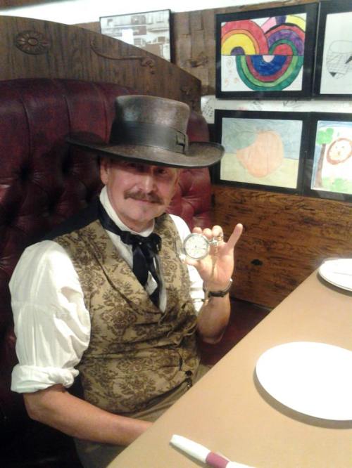 A man in a vintage outfit and hat holds a pocket watch, sitting at a table with empty plates and colorful artwork behind him.