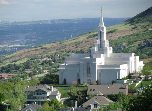 A large white temple with a tall spire, surrounded by green hills and residential buildings in the foreground.