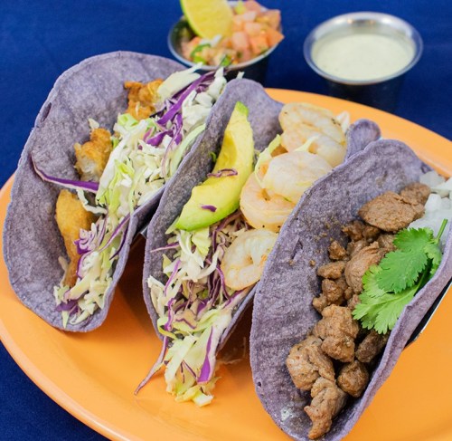 Three colorful tacos on an orange plate: fish, shrimp, and beef, with cabbage and avocado, served with salsa and lime.