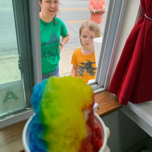 A colorful snow cone in the foreground with a woman and a child looking through a window, both smiling.
