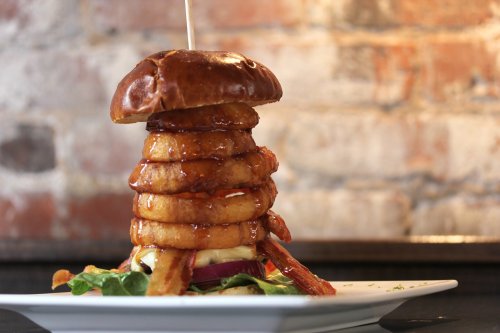 A towering burger with multiple onion rings, drizzled with sauce, served on a plate with greens and a rustic background.