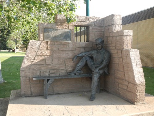 A bronze statue of a seated man in a hat, set against a stone wall with a plaque, in a grassy park area.
