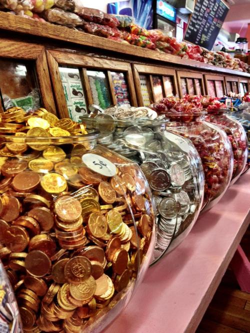 Colorful candy jars filled with gold and silver chocolate coins, lined up on a pink counter in a sweet shop.