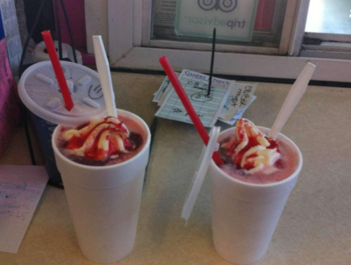 Two large cups with colorful ice cream and syrup, topped with whipped cream and red straws, sit on a table.
