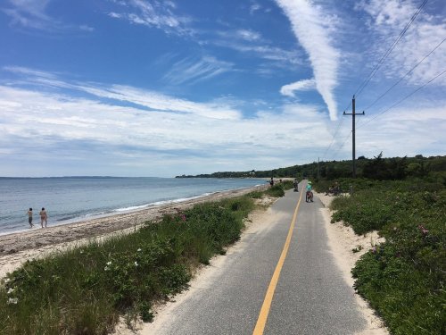 A scenic beachside path with cyclists and walkers, surrounded by greenery under a blue sky with wispy clouds.