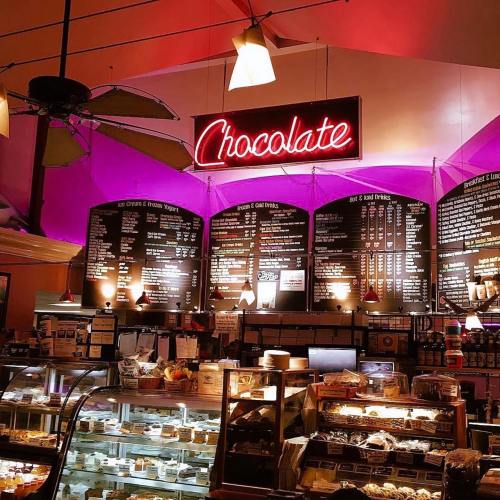 A cozy café interior featuring a neon "Chocolate" sign and a menu board with various dessert options.