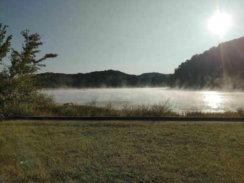 A serene lake at sunrise, with mist rising from the water and hills in the background. Green grass in the foreground.