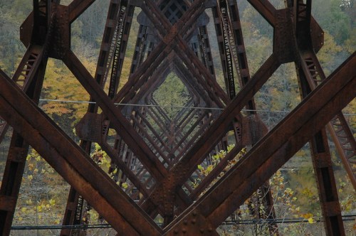 Rusty metal beams create a geometric pattern, framing a view of trees and water in the background.