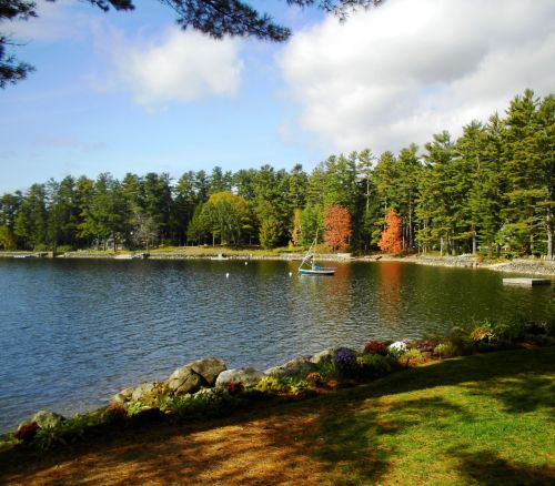 A serene lake surrounded by trees, with autumn colors and a sailboat on the water under a partly cloudy sky.