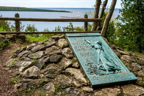 A bronze plaque on a stone mound overlooks a serene lake, surrounded by trees and a wooden fence.