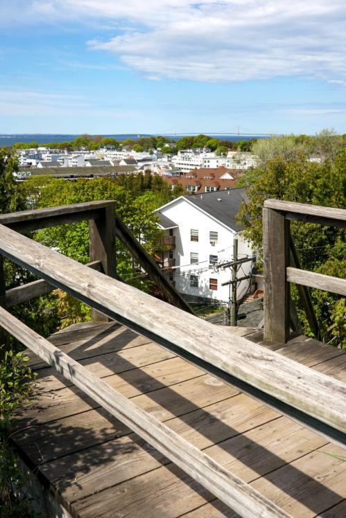 A wooden walkway leads to a scenic view of a town and distant water under a partly cloudy sky.