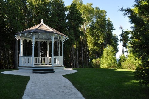 A white gazebo surrounded by lush green trees and a sandy path leading to it.