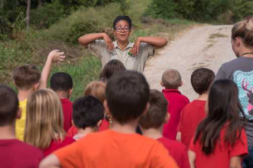 A group of children in colorful shirts listen to an instructor on a nature trail, engaging in a fun activity.