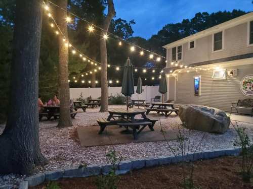 Outdoor seating area with picnic tables, string lights, and trees, creating a cozy atmosphere at dusk.