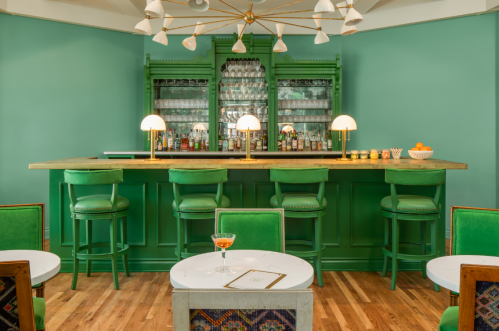 A stylish bar with a green backdrop, featuring a wooden counter, green stools, and a cocktail on a white table.