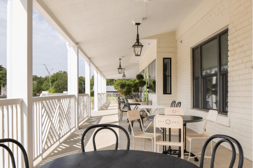 A spacious porch with black tables and chairs, featuring hanging lanterns and greenery, under a bright sky.