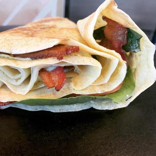 A close-up of a folded tortilla wrap filled with bacon and leafy greens, resting on a dark surface.