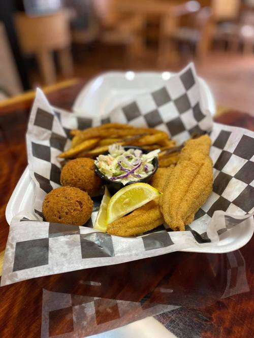A takeout tray with fried fish, coleslaw, lemon wedges, sweet potato fries, and two hushpuppies on a checkered paper.