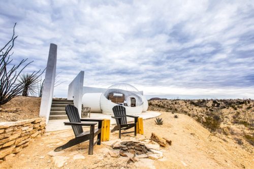 A unique bubble-shaped structure in a desert landscape, with two chairs and a cloudy sky above.