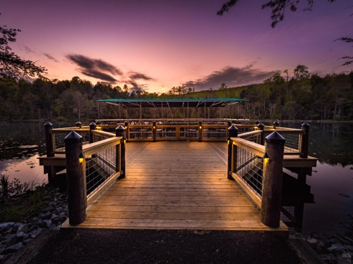 A wooden dock extends over a calm lake at sunset, surrounded by trees and softly lit by lanterns.