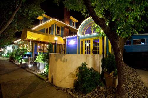 A brightly lit restaurant at night, featuring colorful architecture and lush greenery along the sidewalk.