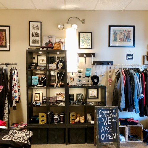 A cozy boutique interior with clothing racks, jewelry displays, and a sign that says "Beyond We're Open."
