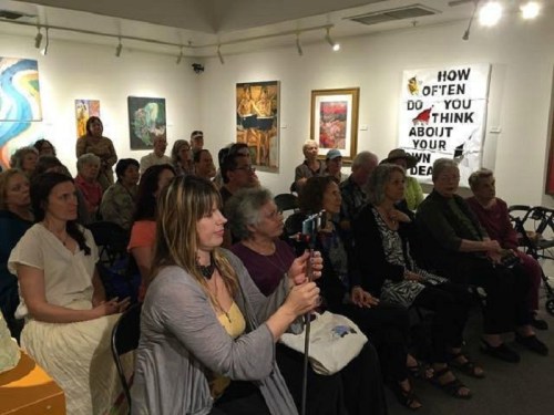 A diverse audience seated in an art gallery, engaged in a presentation, with various artworks displayed on the walls.