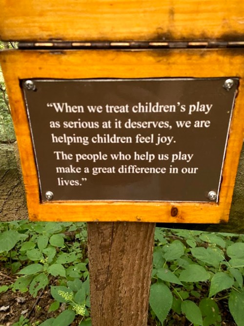 A wooden sign in a forest with a quote about the importance of treating children's play seriously for their joy.