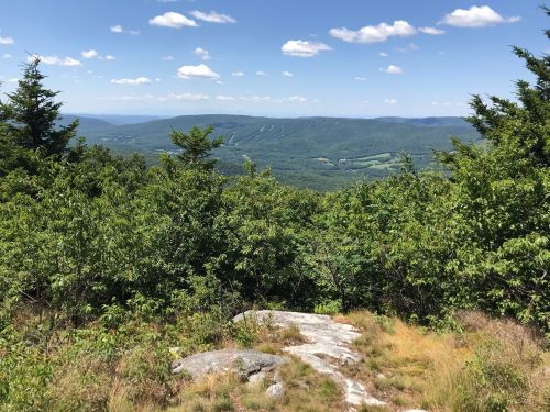 A scenic view from a rocky overlook, showcasing rolling green hills and a blue sky with scattered clouds.