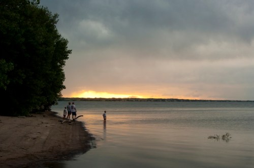 A serene beach scene at sunset with three people walking along the shore and one child wading in the water.