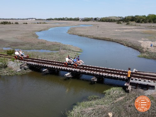 A group of people rides on a rail cart over a wooden bridge, surrounded by marshland and a winding river.