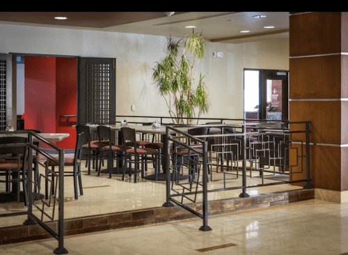 Interior of a restaurant featuring tables, chairs, and a decorative railing, with a plant in the background.