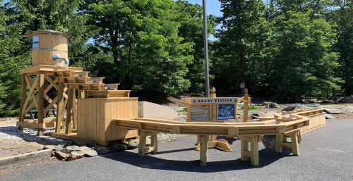 A wooden assay station with a water feature, steps, and a sign, surrounded by trees in a sunny outdoor setting.