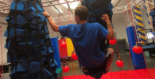 A person in a blue shirt climbs a blue foam obstacle in an indoor adventure course, surrounded by colorful equipment.