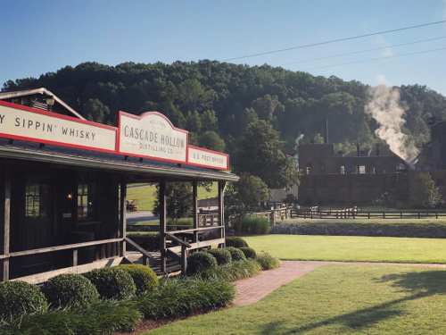 A distillery building with a porch, surrounded by green hills and smoke rising from a nearby structure.