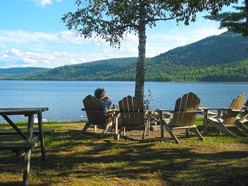 A person sits in an Adirondack chair by a lake, surrounded by trees and mountains under a blue sky.