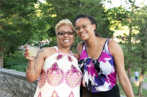 Two women smile together outdoors, one holding a drink, surrounded by greenery.