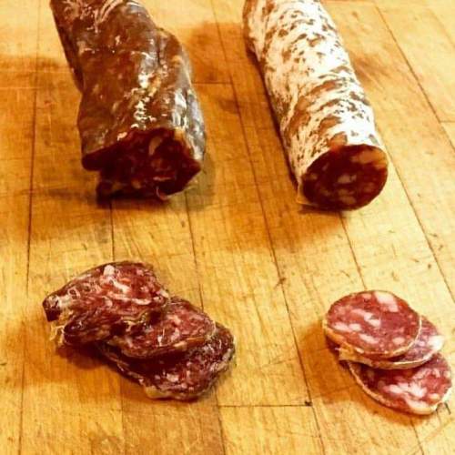 Three types of cured meats displayed on a wooden surface, including whole logs and sliced pieces.