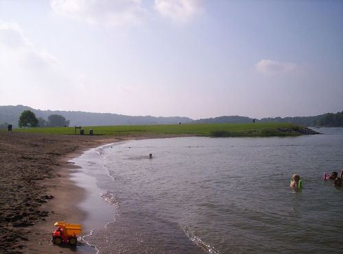A sandy beach by a calm river, with children playing in the water and a toy truck on the shore.