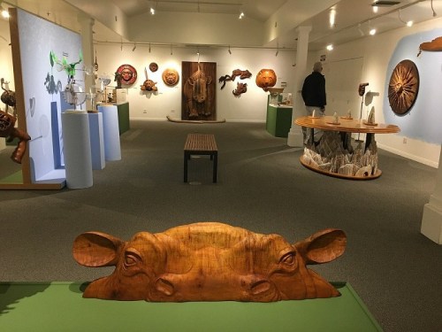 Art gallery featuring wooden sculptures and masks, with a wooden cow head in the foreground and visitors exploring the space.
