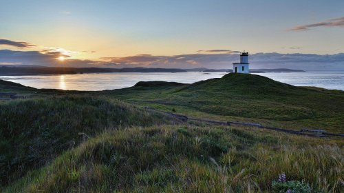 A serene landscape featuring a lighthouse on a grassy hill at sunset, overlooking calm waters and distant islands.