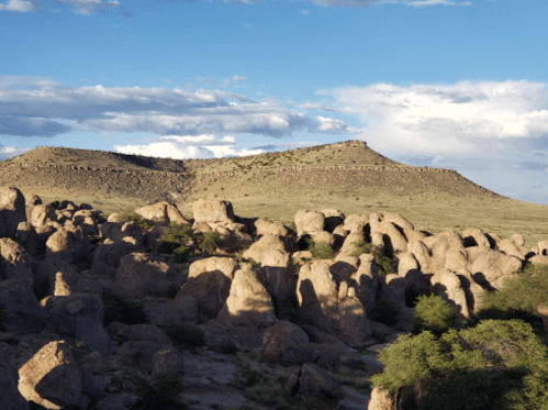 A rocky landscape with large boulders and a distant hill under a blue sky with scattered clouds.